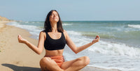 Jeune femme pastiquant la méditation face au soleil assise sur la plage en posture tailleur
