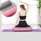 Femme assise de dos sur le traversin de yoga adhocia éco de suède les jambes croisées, le bras droit en l'air, la main gauche en appui sur le traversin de couleur rose.